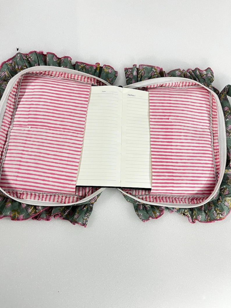 May include: An open notebook inside a floral print case with a pink and white striped lining. The case has a ruffled edge and a zip closure. The notebook is open to a blank page with the word "Notes" visible.