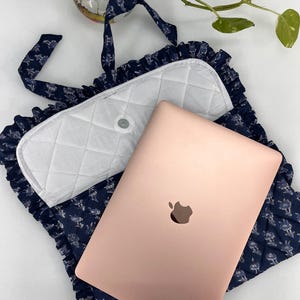 May include: A rose gold laptop rests on a navy blue quilted fabric sleeve featuring a white floral pattern and ruffled edges. The sleeve has a white quilted flap with a central snap closure and matching navy blue fabric straps for carrying.