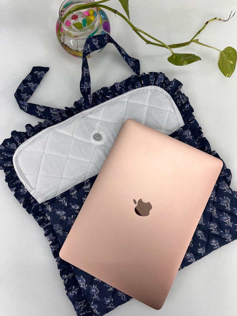 May include: A rose gold laptop rests on a navy blue quilted fabric sleeve featuring a white floral pattern and ruffled edges. The sleeve has a white quilted flap with a central snap closure and matching navy blue fabric straps for carrying.