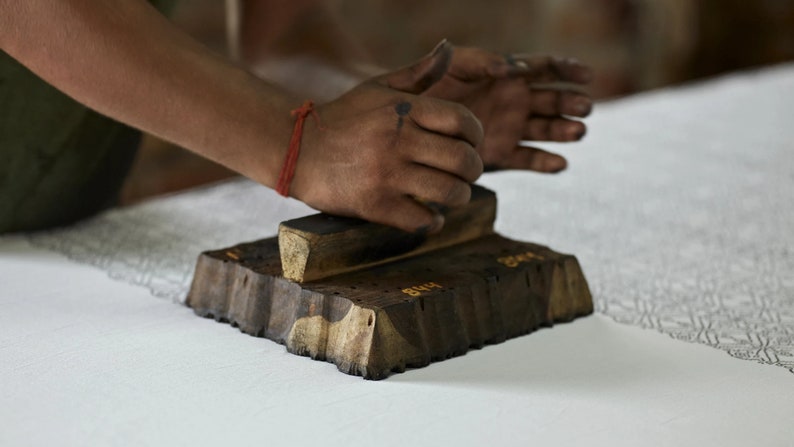 Puede incluir: Una persona utilizando un sello de impresi&oacute;n de madera para crear un patr&oacute;n en una tela blanca. El sello es de color marr&oacute;n oscuro con un mango de color marr&oacute;n claro y tiene un dise&ntilde;o detallado tallado.