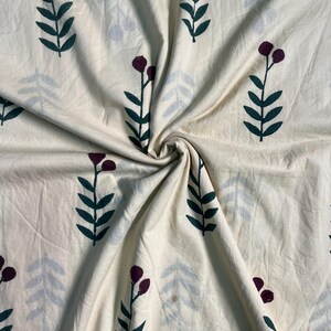 May include: Cream-colored fabric with a repeating pattern of dark green leaves and burgundy berry clusters on stems. Faint blue-gray leaf shapes are also scattered across the fabric. The fabric is gathered and folded, showing its texture.