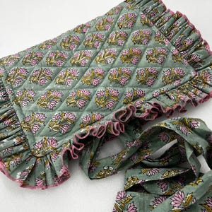 May include: A quilted, sage green fabric item with a floral pattern in pink and yellow. The edges are trimmed with a ruffled border in the same green, accented with pink. A matching fabric tie is also visible.