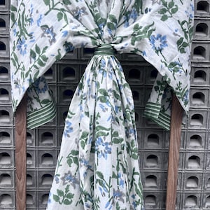 K&ouml;nnte beinhalten: Ein wei&szlig;er Schal mit blau-gr&uuml;nen Blumenmustern. Der Schal ist &uuml;ber einem Holzrahmen drapiert, der das Design hervorhebt. Die Kanten sind mit einem gr&uuml;nen Rand versehen.