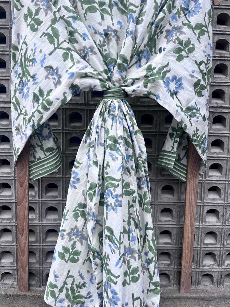 K&ouml;nnte beinhalten: Ein wei&szlig;er Schal mit blau-gr&uuml;nen Blumenmustern. Der Schal ist &uuml;ber einem Holzrahmen drapiert, der das Design hervorhebt. Die Kanten sind mit einem gr&uuml;nen Rand versehen.