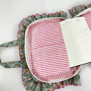 May include: A notebook case with a floral print, white zip, and pink striped lining. The case has a ruffled edge and a matching handle. An open notebook with lined pages is visible.