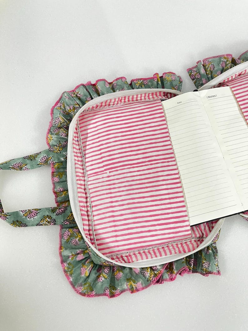 May include: A notebook case with a floral print, white zip, and pink striped lining. The case has a ruffled edge and a matching handle. An open notebook with lined pages is visible.