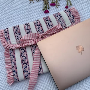 Puede incluir: Una funda para portátil a rayas rosas y blancas con un borde con volantes y una cinta a juego. La funda presenta un estampado floral en azul y rosa. Un portátil de oro rosa es parcialmente visible.
