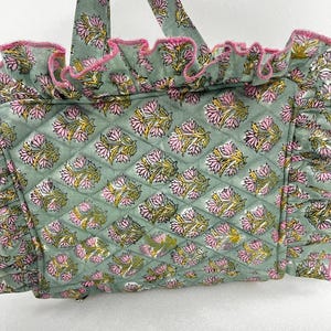 May include: A quilted tote bag in a soft green hue, decorated with a repeating floral pattern in pink and yellow. The bag has ruffled edges trimmed with pink and matching handles. The design blends functionality with floral aesthetics.
