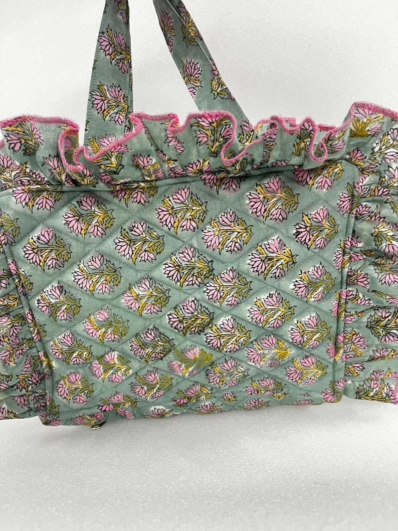 May include: A quilted tote bag in a soft green hue, decorated with a repeating floral pattern in pink and yellow. The bag has ruffled edges trimmed with pink and matching handles. The design blends functionality with floral aesthetics.