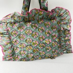 May include: A quilted tote bag with a floral pattern in shades of green, pink, and yellow. The bag has a ruffled edge with pink trim and matching handles. The bag features a zip closure.
