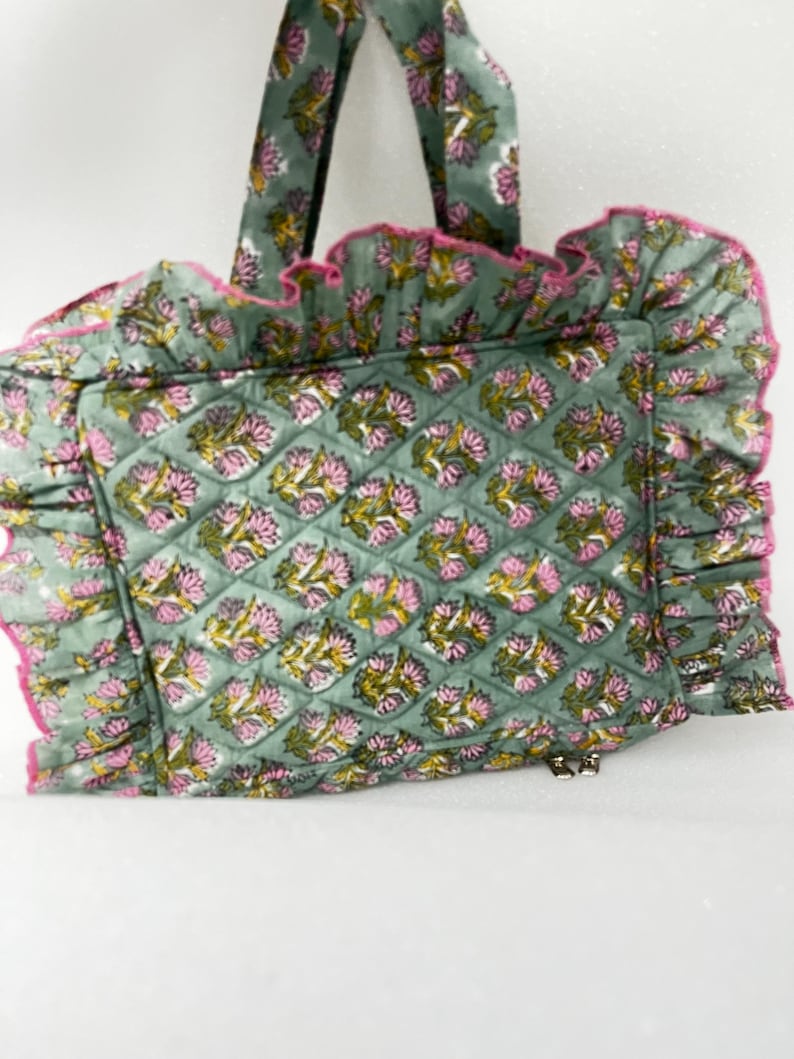 May include: A quilted tote bag with a floral pattern in shades of green, pink, and yellow. The bag has a ruffled edge with pink trim and matching handles. The bag features a zip closure.