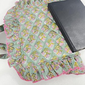 May include: A sage green quilted tote bag with a floral pattern in pink and yellow. The bag features a ruffled edge with pink trim. A black book rests on top.