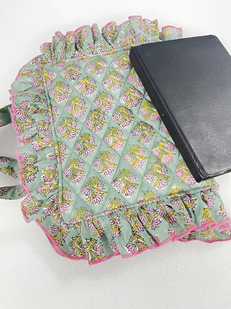 May include: A sage green quilted tote bag with a floral pattern in pink and yellow. The bag features a ruffled edge with pink trim. A black book rests on top.