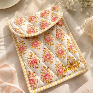 May include: A quilted pouch in off-white, with a flap closure. It displays a repeating floral pattern in pink and yellow, and a yellow and white striped border. The design is accented with a gold embroidered detail.