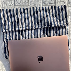 Op de afbeelding: Een roségouden laptop met een zwart Apple-logo ligt op een gewatteerde laptoptas met marineblauwe en witte strepen. De hoes is omgevouwen en het patroon bestaat uit verticale strepen. De achtergrond is een wit, getextureerd oppervlak.