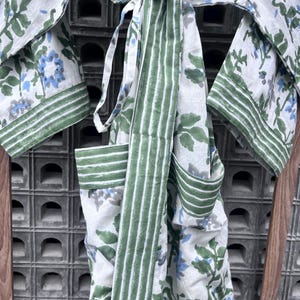 K&ouml;nnte beinhalten: Ein wei&szlig;es Bademantel mit floralem Muster und gr&uuml;nen und blauen Akzenten. Der Bademantel hat einen gestreiften gr&uuml;nen G&uuml;rtel und Besatz. Die &Auml;rmel und Taschen sind sichtbar, und der Bademantel ist an der Taille gebunden. Das Blumenmuster umfasst gr&uuml;ne Bl&auml;tter und blaue Bl&uuml;ten.