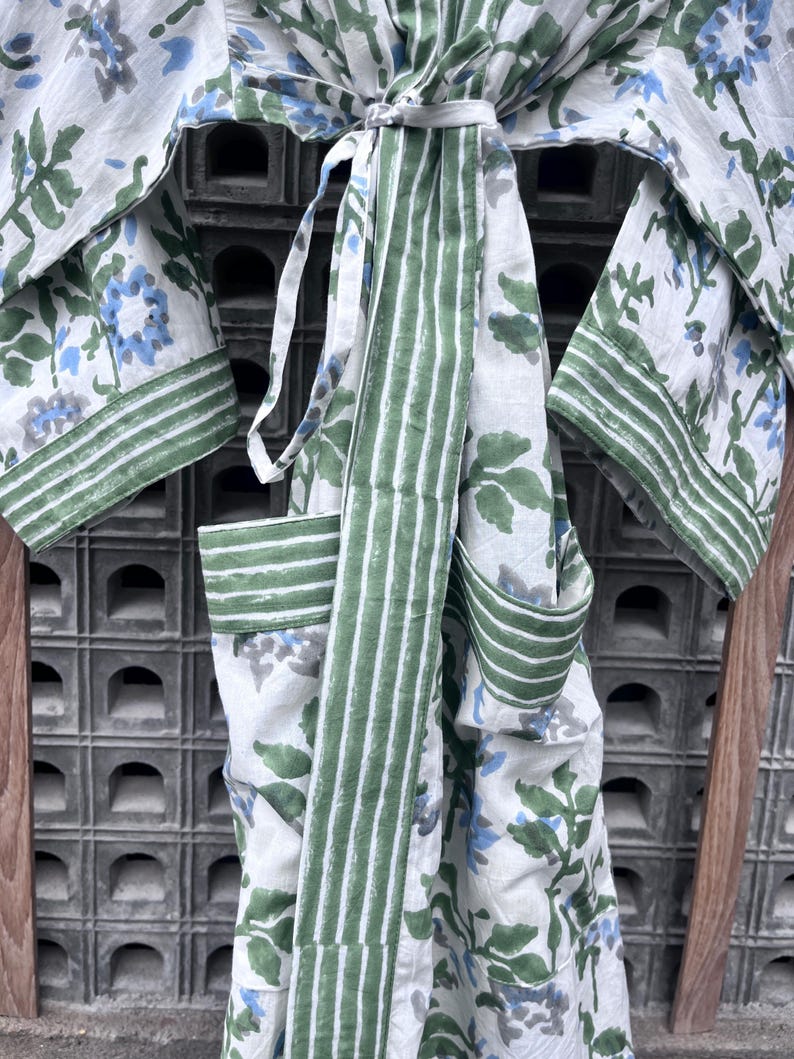 K&ouml;nnte beinhalten: Ein wei&szlig;es Bademantel mit floralem Muster und gr&uuml;nen und blauen Akzenten. Der Bademantel hat einen gestreiften gr&uuml;nen G&uuml;rtel und Besatz. Die &Auml;rmel und Taschen sind sichtbar, und der Bademantel ist an der Taille gebunden. Das Blumenmuster umfasst gr&uuml;ne Bl&auml;tter und blaue Bl&uuml;ten.