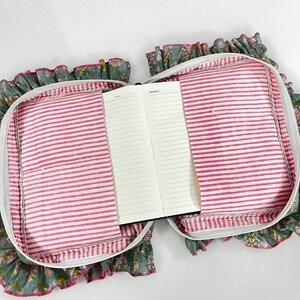 May include: An open notebook with lined pages is placed inside a floral print case. The case has a pink and white striped lining, a white zip, and a ruffled edge with a floral pattern in green, pink, and white.