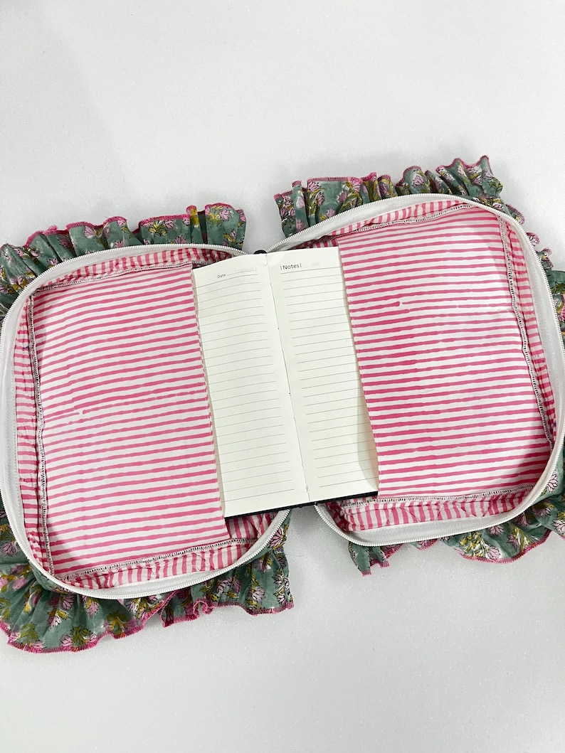 May include: An open notebook with lined pages is placed inside a floral print case. The case has a pink and white striped lining, a white zip, and a ruffled edge with a floral pattern in green, pink, and white.