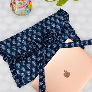 May include: A quilted navy blue laptop sleeve featuring a white floral pattern, a ruffled edge, and a fabric tie closure, positioned next to a rose gold laptop with a visible apple logo on the lid, providing a protective cover for the device.