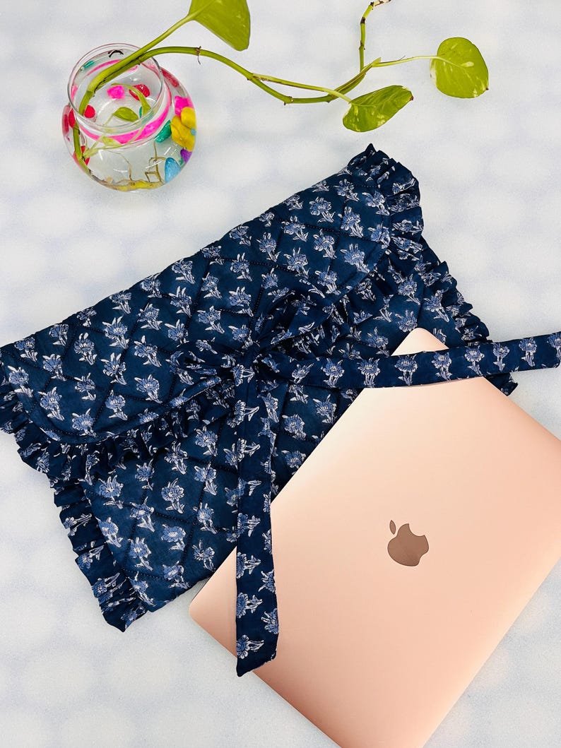 May include: A quilted navy blue laptop sleeve featuring a white floral pattern, a ruffled edge, and a fabric tie closure, positioned next to a rose gold laptop with a visible apple logo on the lid, providing a protective cover for the device.