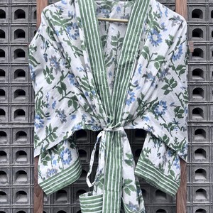 K&ouml;nnte beinhalten: Wei&szlig;er Bademantel mit floralem Muster, gr&uuml;nen Zierleisten und passendem Bindeband. Der Bademantel hat ein Muster aus blauen und grauen Blumen mit gr&uuml;nen Bl&auml;ttern. Der Bademantel h&auml;ngt an einem Holzb&uuml;gel vor einem strukturierten Hintergrund.