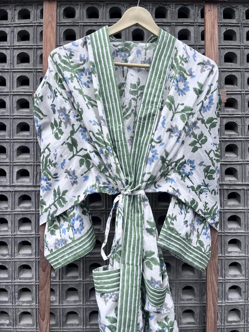 K&ouml;nnte beinhalten: Wei&szlig;er Bademantel mit floralem Muster, gr&uuml;nen Zierleisten und passendem Bindeband. Der Bademantel hat ein Muster aus blauen und grauen Blumen mit gr&uuml;nen Bl&auml;ttern. Der Bademantel h&auml;ngt an einem Holzb&uuml;gel vor einem strukturierten Hintergrund.