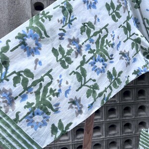 K&ouml;nnte beinhalten: Wei&szlig;er Stoff mit einem Blumenmuster mit blauen und grauen Blumen, gr&uuml;nen Bl&auml;ttern und Stielen. Der Rand des Stoffes hat einen gr&uuml;n-wei&szlig; gestreiften Rand. Der Stoff scheint leicht und f&uuml;r Kleidung oder Heimdekoration geeignet zu sein.