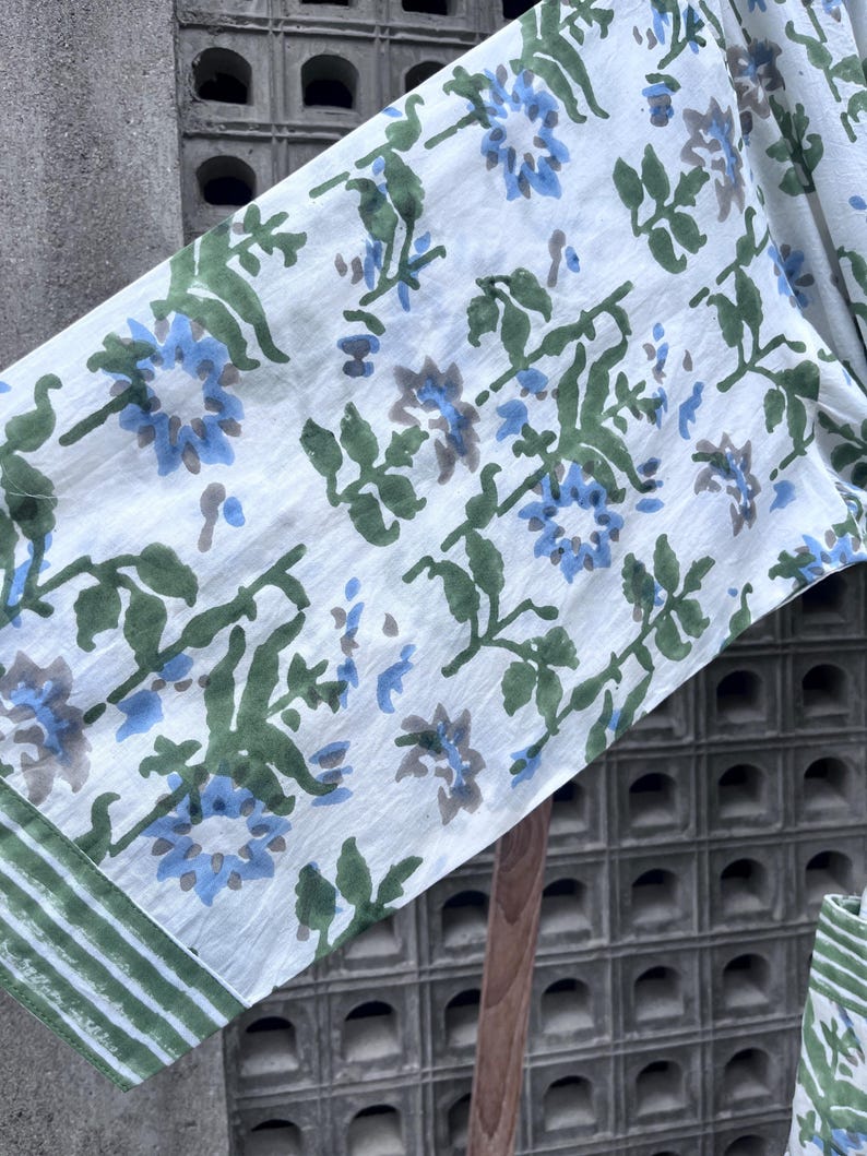 K&ouml;nnte beinhalten: Wei&szlig;er Stoff mit einem Blumenmuster mit blauen und grauen Blumen, gr&uuml;nen Bl&auml;ttern und Stielen. Der Rand des Stoffes hat einen gr&uuml;n-wei&szlig; gestreiften Rand. Der Stoff scheint leicht und f&uuml;r Kleidung oder Heimdekoration geeignet zu sein.