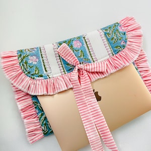 May include: A padded laptop sleeve featuring a floral pattern in blue, green, and white, accented with pink and white striped ruffled edges and a matching fabric tie closure. The sleeve partially encases a gold laptop with a visible apple logo.