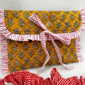 May include: A quilted clutch with a mustard yellow floral pattern and pink and white striped ruffle trim. The clutch is tied with a matching pink and white striped ribbon. A separate red and white striped ruffle is also visible.