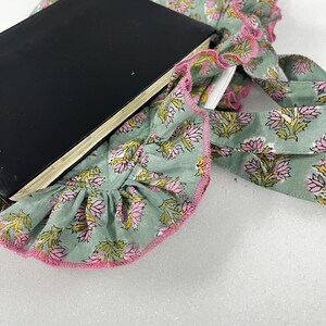 May include: A black notebook lies on a light green fabric with a floral pattern in pink and yellow. The fabric has a ruffled edge trimmed with pink. The notebook is open, revealing white pages.
