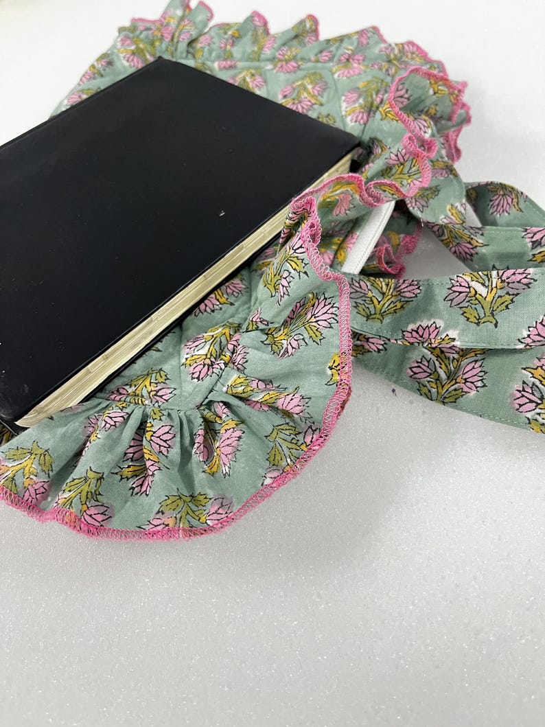 May include: A black notebook lies on a light green fabric with a floral pattern in pink and yellow. The fabric has a ruffled edge trimmed with pink. The notebook is open, revealing white pages.