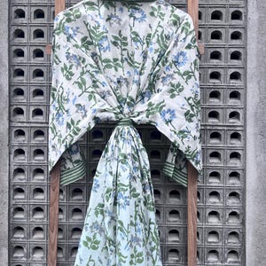 K&ouml;nnte beinhalten: Ein wei&szlig;er Bademantel mit einem Blumenmuster aus blauen Blumen und gr&uuml;nen Bl&auml;ttern, der an einem Holzgestell h&auml;ngt. Der Bademantel hat gr&uuml;ne Verzierungen an Kragen und Manschetten. Der Hintergrund ist eine graue, strukturierte Wand.