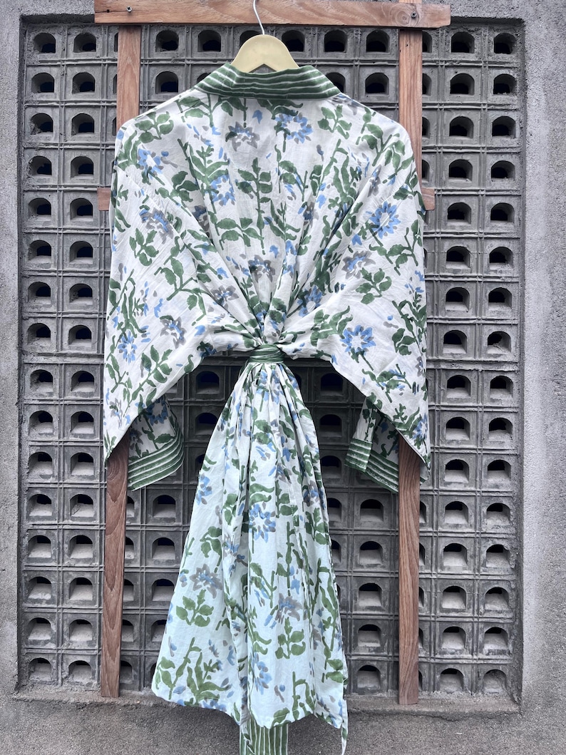 K&ouml;nnte beinhalten: Ein wei&szlig;er Bademantel mit einem Blumenmuster aus blauen Blumen und gr&uuml;nen Bl&auml;ttern, der an einem Holzgestell h&auml;ngt. Der Bademantel hat gr&uuml;ne Verzierungen an Kragen und Manschetten. Der Hintergrund ist eine graue, strukturierte Wand.