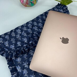 May include: A rose gold laptop rests on a dark blue quilted fabric sleeve featuring a white floral pattern and ruffled edges. The sleeve has a flap closure and a tie detail. A small glass vase with colorful stones and a green plant stem is visible.