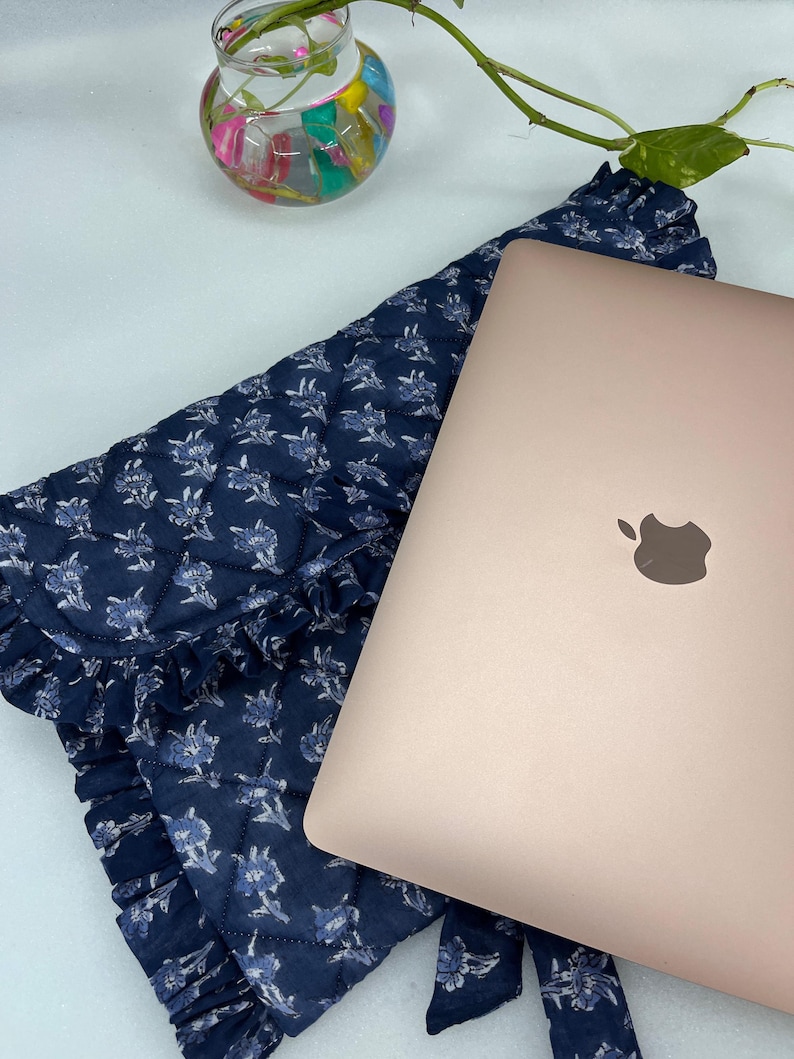 May include: A rose gold laptop rests on a dark blue quilted fabric sleeve featuring a white floral pattern and ruffled edges. The sleeve has a flap closure and a tie detail. A small glass vase with colorful stones and a green plant stem is visible.