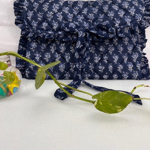 May include: A navy blue quilted fabric pouch featuring a white floral pattern, a ruffled edge trim, and a tie closure. The rectangular pouch has a diamond-stitched texture and is positioned behind a small glass vase containing a green plant.