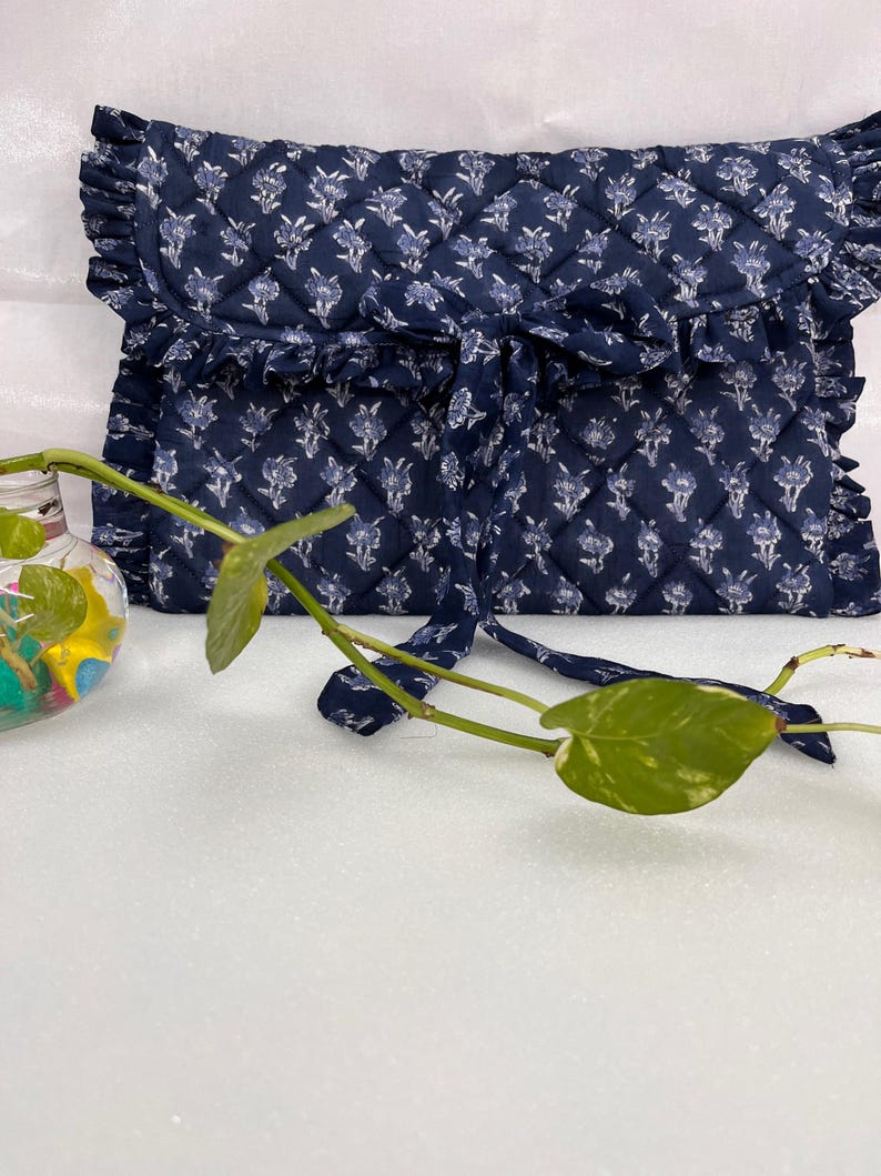 May include: A navy blue quilted fabric pouch featuring a white floral pattern, a ruffled edge trim, and a tie closure. The rectangular pouch has a diamond-stitched texture and is positioned behind a small glass vase containing a green plant.