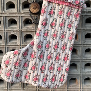 Könnte beinhalten: Ein weißer, gesteppter Weihnachtssocken mit einem rosa-weiß gestreiften Rüschenbesatz oben. Der Socken ist mit einem rosa Blumendesign verziert.