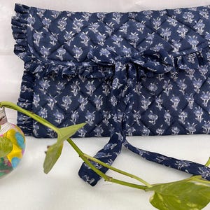 May include: A navy blue quilted clutch with a white floral pattern, featuring a ruffled edge along the flap and a matching fabric tie closure. The rectangular bag has a diamond-stitched texture and a soft, fabric construction throughout.