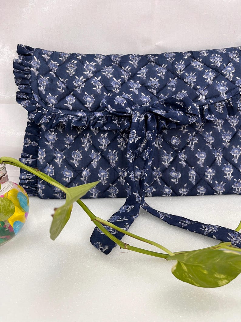 May include: A navy blue quilted clutch with a white floral pattern, featuring a ruffled edge along the flap and a matching fabric tie closure. The rectangular bag has a diamond-stitched texture and a soft, fabric construction throughout.