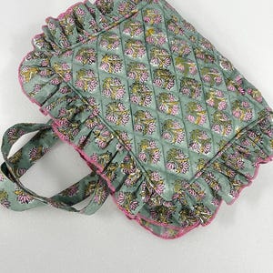 May include: A quilted tote bag with a floral pattern in pink, yellow, and green. The bag has a ruffled edge with pink trim and matching handles. The bag is approximately 35 cm wide and 30 cm tall.