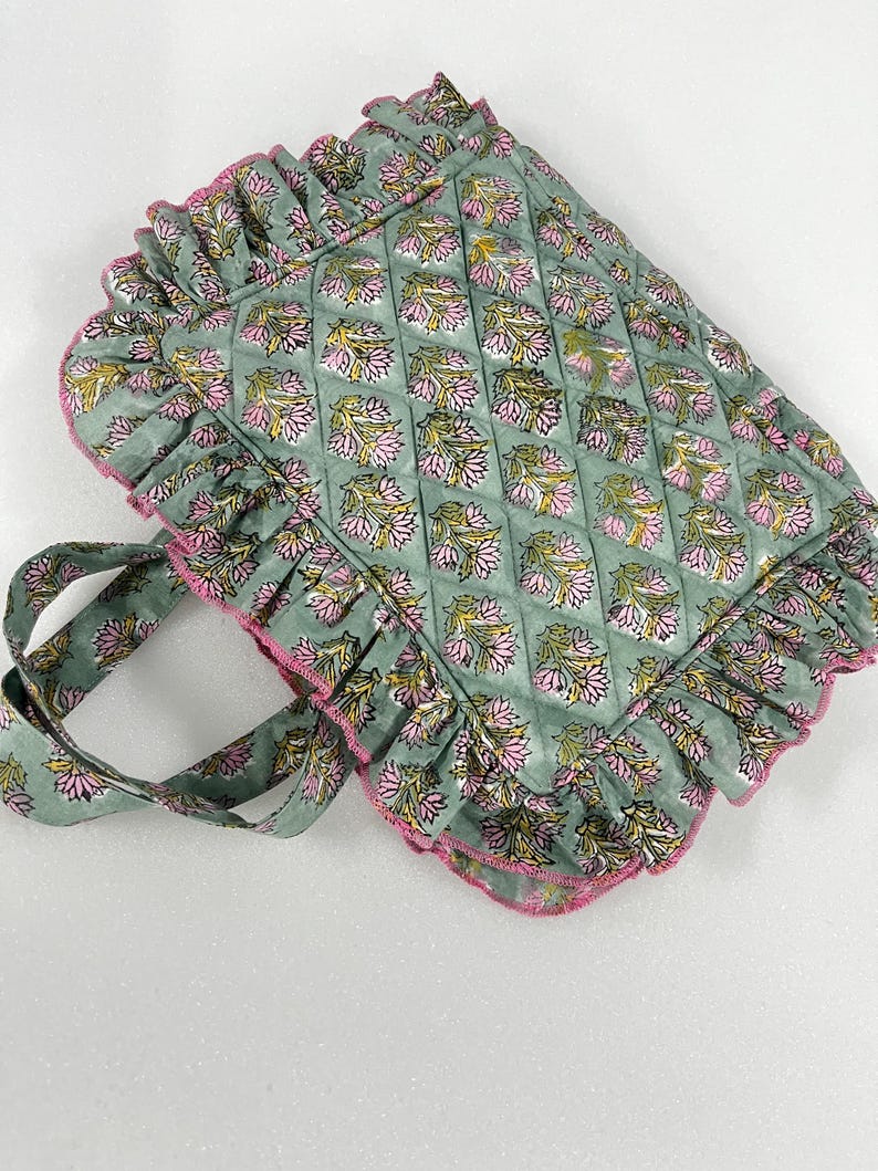May include: A quilted tote bag with a floral pattern in pink, yellow, and green. The bag has a ruffled edge with pink trim and matching handles. The bag is approximately 35 cm wide and 30 cm tall.