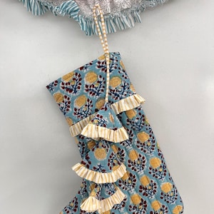 May include: A blue Christmas stocking with a floral pattern in yellow and maroon. The stocking has ruffled trim in yellow and white stripes. A matching striped loop is attached for hanging. A ruffled garland with red and white stripes is at the top.