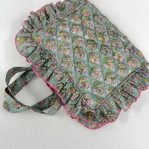 Puede incluir: Un bolso tote acolchado con un estampado floral en rosa y amarillo sobre un fondo verde salvia. El bolso tiene un borde con volantes con ribete rosa y asas a juego. El bolso está diseñado para transportar objetos.
