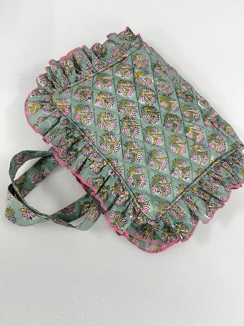 May include: A sage green quilted tote bag featuring a floral pattern in pink and yellow. The bag has a ruffled edge with pink trim and matching handles. The bag is designed for carrying items.