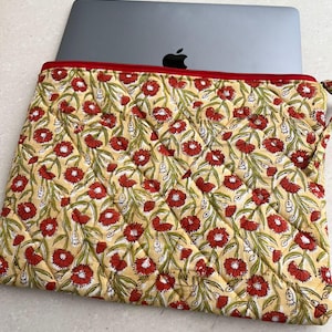 Puede incluir: Una funda para portátil amarilla con una cremallera roja y un estampado floral con flores rojas y hojas verdes.