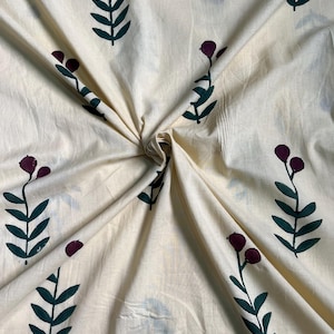 May include: Cream-colored fabric with a repeating pattern of stylized floral elements. The design features dark green leaves and burgundy-colored berry-like shapes. The fabric is folded, creating shadows and texture.