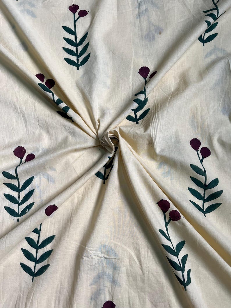 May include: Cream-colored fabric with a repeating pattern of stylized floral elements. The design features dark green leaves and burgundy-colored berry-like shapes. The fabric is folded, creating shadows and texture.